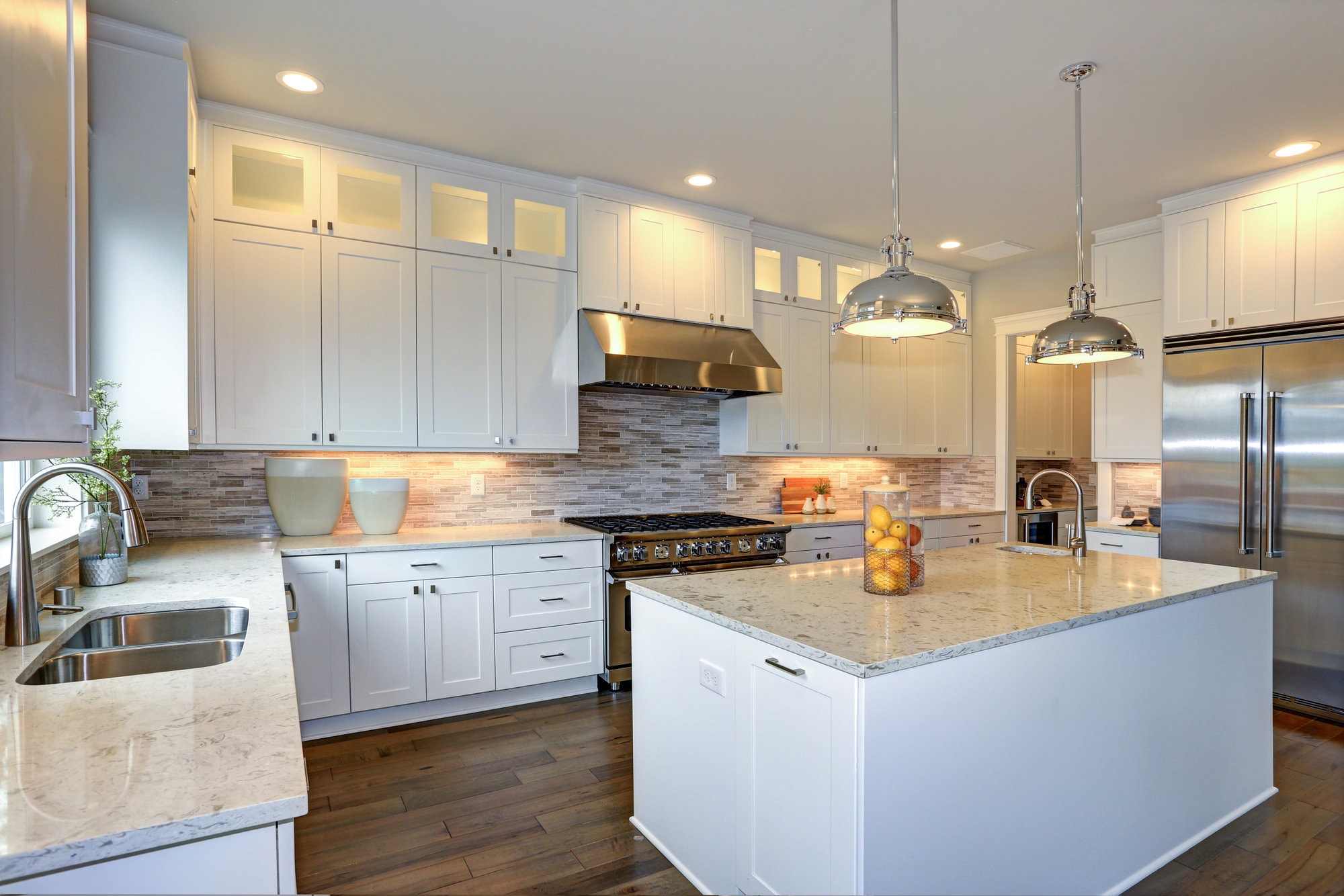 Amazing white kitchen design with white shaker cabinets paired with  white and gray marble counters, large White kitchen peninsula with a sink and hardwood kitchen floors.