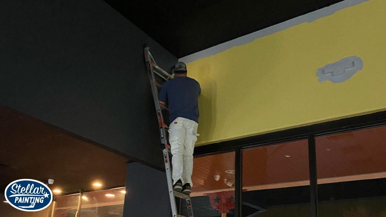 Commercial Painting Alexandra Meadows Fort Worth, TX - Contract Types - Stellar Painting This is a commercial interior painting project by Stellar Painting in Alexandra Meadows Fort Worth, TX, showing a painter applying bright yellow paint above a storefront entrance.